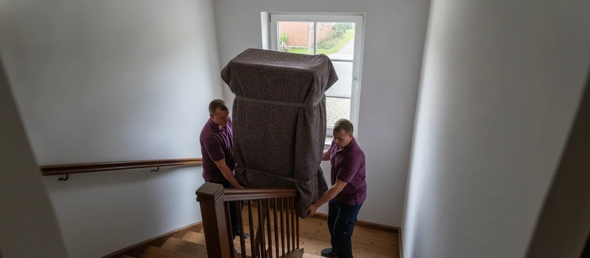 Zwei Umzugshelfer tragen einen in Decken verpackten Schrank über ein enges Treppenpodest